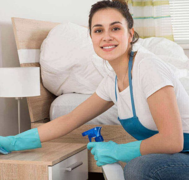 A woman cleaning a bedside table with a smile.