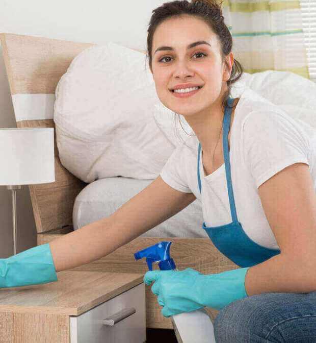 A woman cleaning a bedside table with a smile.