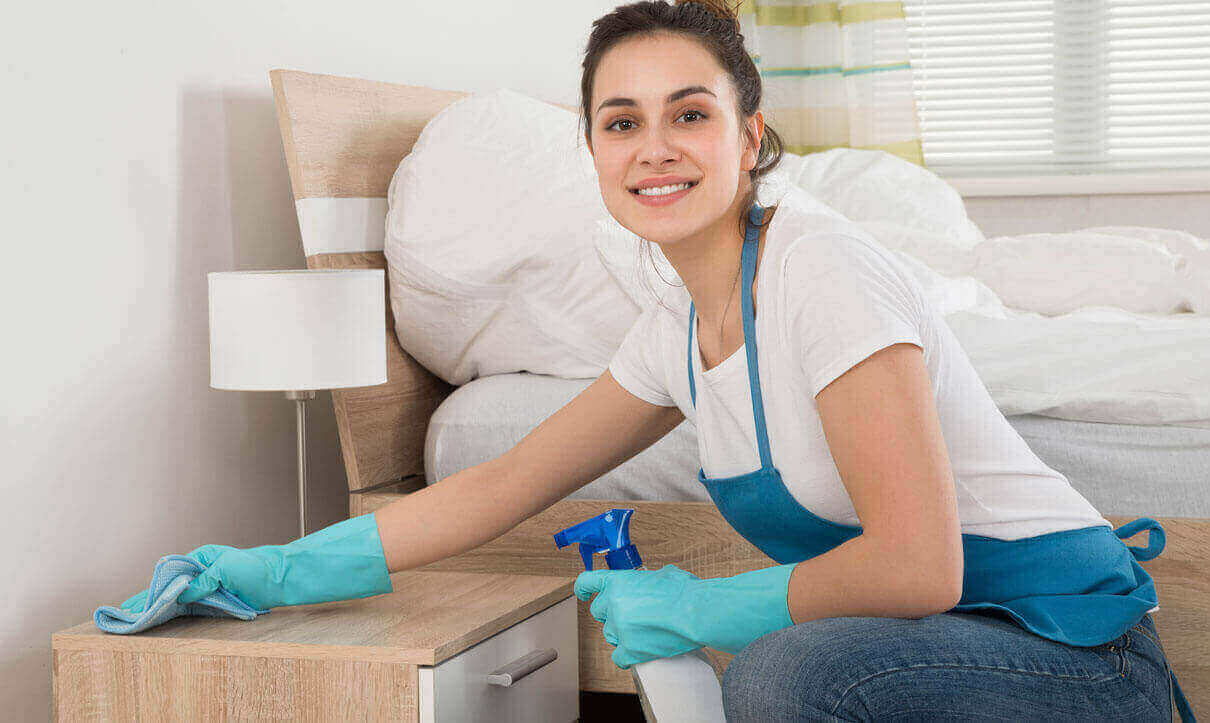 A woman cleaning a bedside table with a smile.